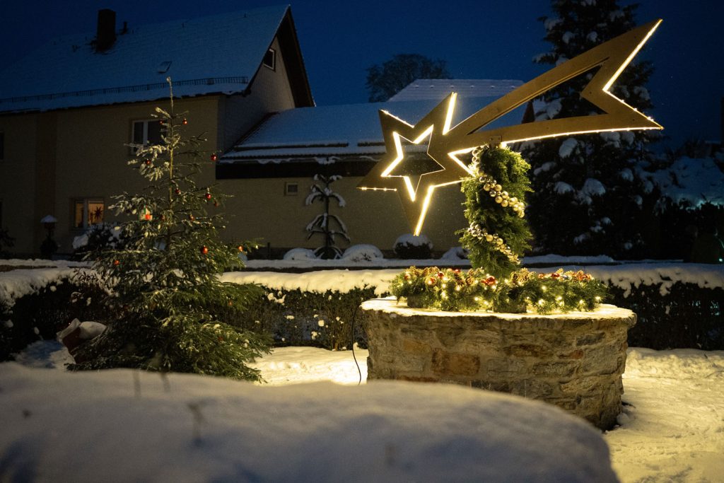 Weihnachtlich geschmückter Dorfbrunnen im Schnee, mit leuchtender Sternschnuppe aus Holz und Tannenzweigen, daneben ein Weihnachtsbaum. 