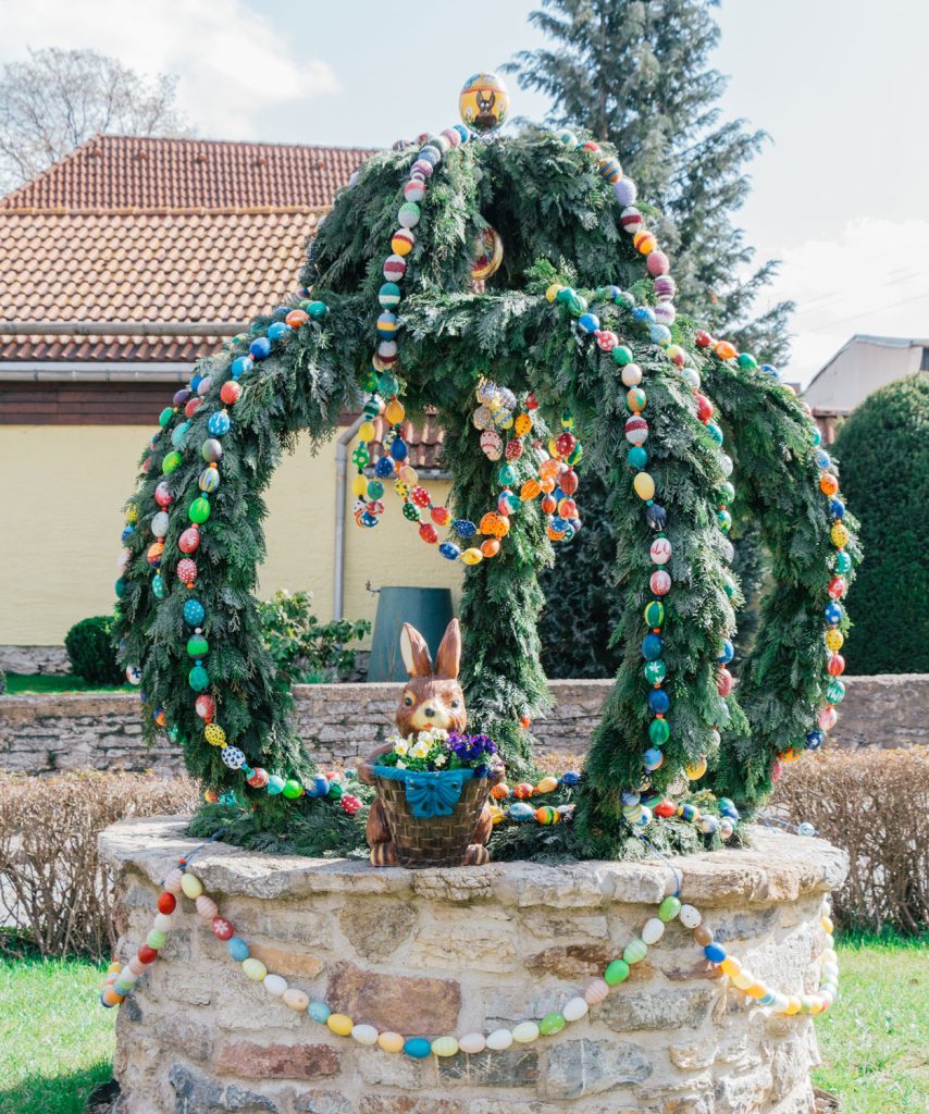 Geschmückte Krone aus Tannenzweigen und bunten Ostereiern auf dem Löhmaer Dorfbrunnen, mit Osterhasenfigur im Vordergrund