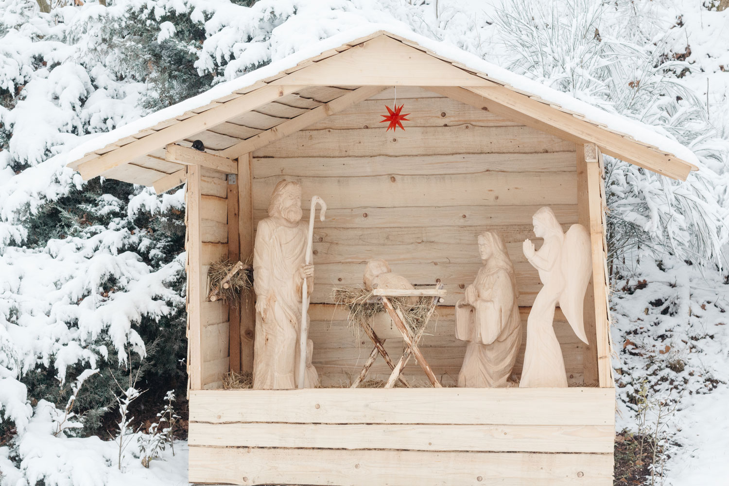 Eine hölzerne Krippe steht im Schnee, umgeben von einer winterlichen Landschaft und sanften Schneeflocken.